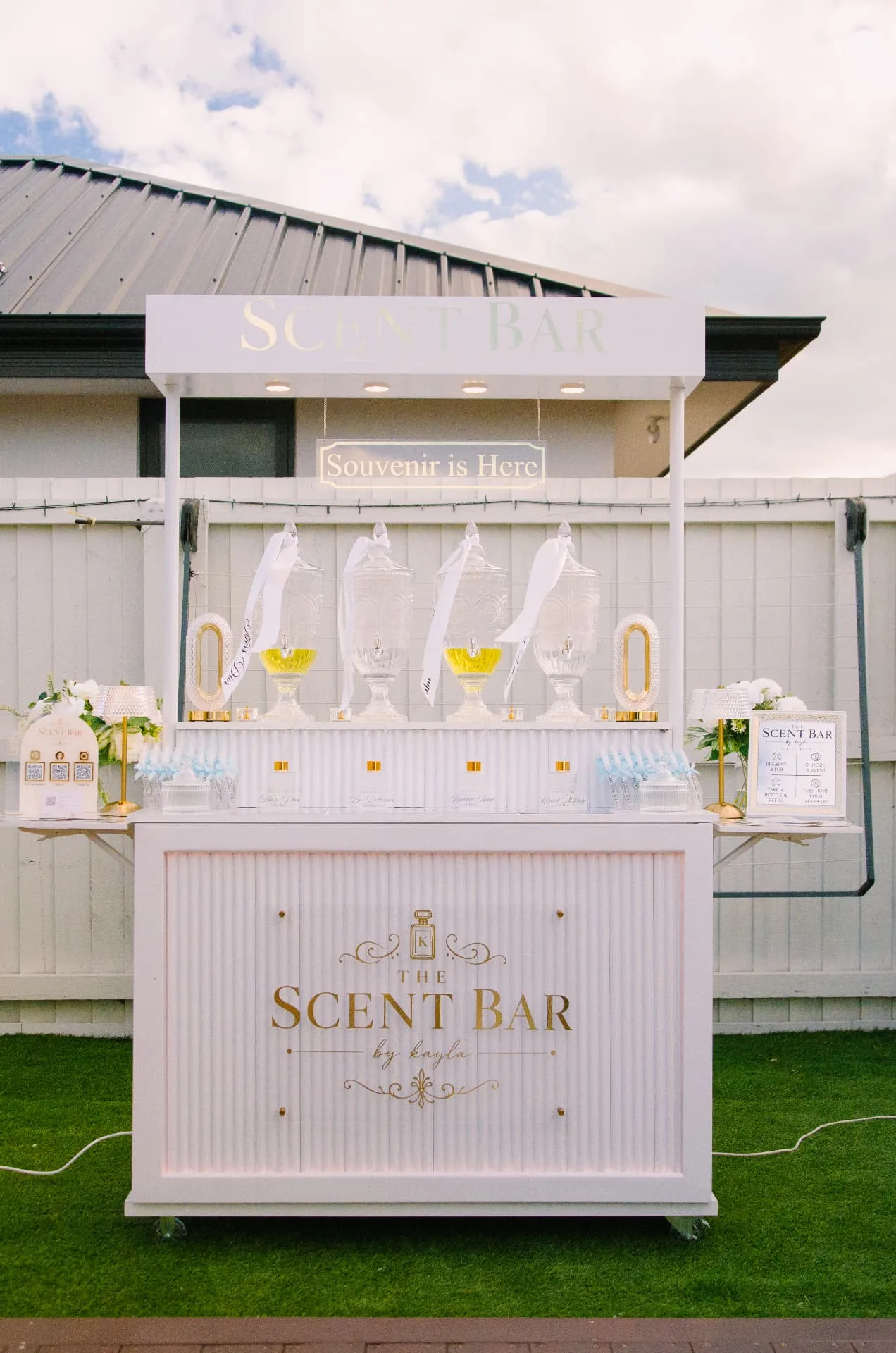 The Scent Bar by Kayla white mobile perfume bar with the Souvenir is Here sign and crystal fragrance dispensers at a garden event